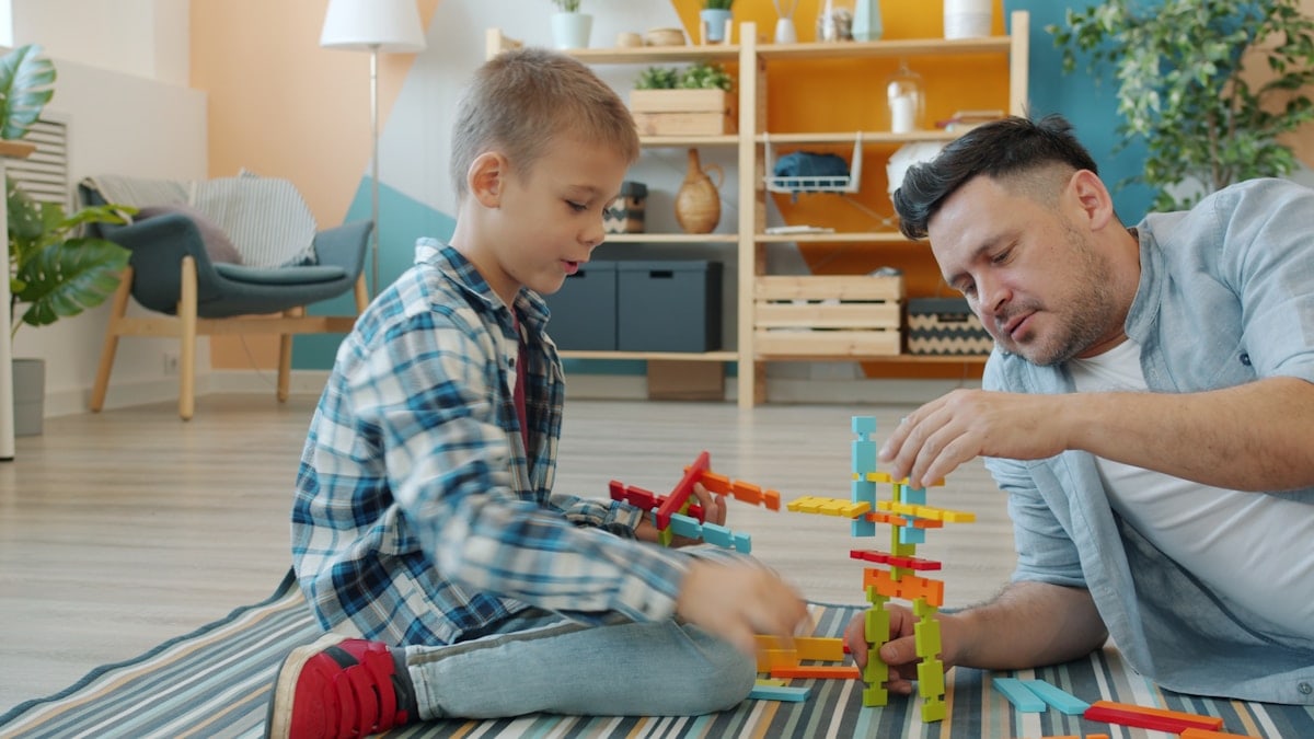 Happy kid is playing with joyful dad using construction blocks enjoying funny game at home on floor having fun and talking. Childhood and people concept.